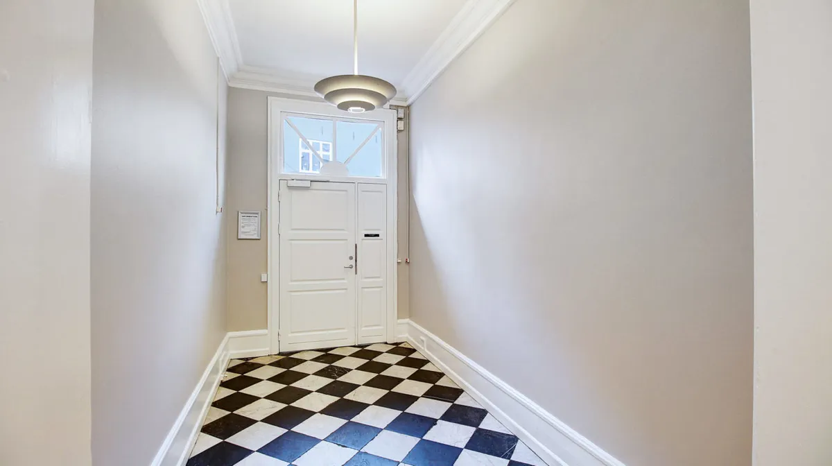 Et halvmørkt korridor med sort og hvid tæppegulv og hvide vægge, med en åben hvid dør og et hvidt glasvindue i baggrunden