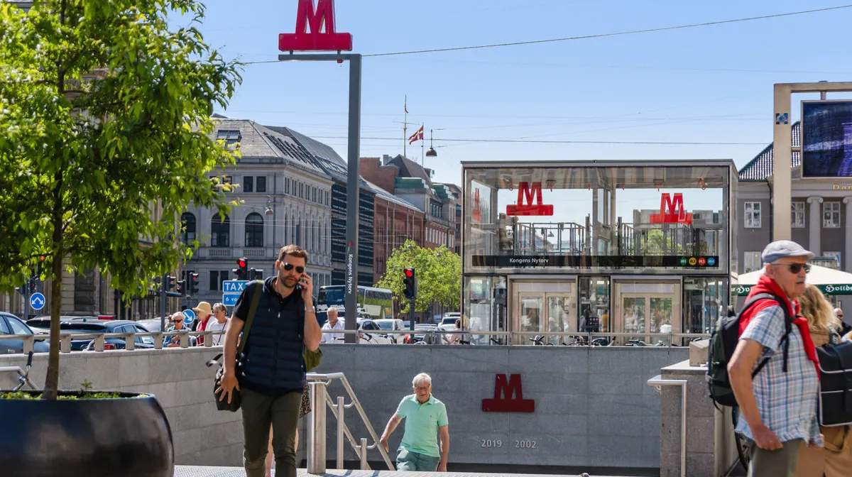 En mand taler på mobiltelefonen og går ned ad trappen i Kongens Nytorv Metrostation med mange mennesker og biler i baggrunden