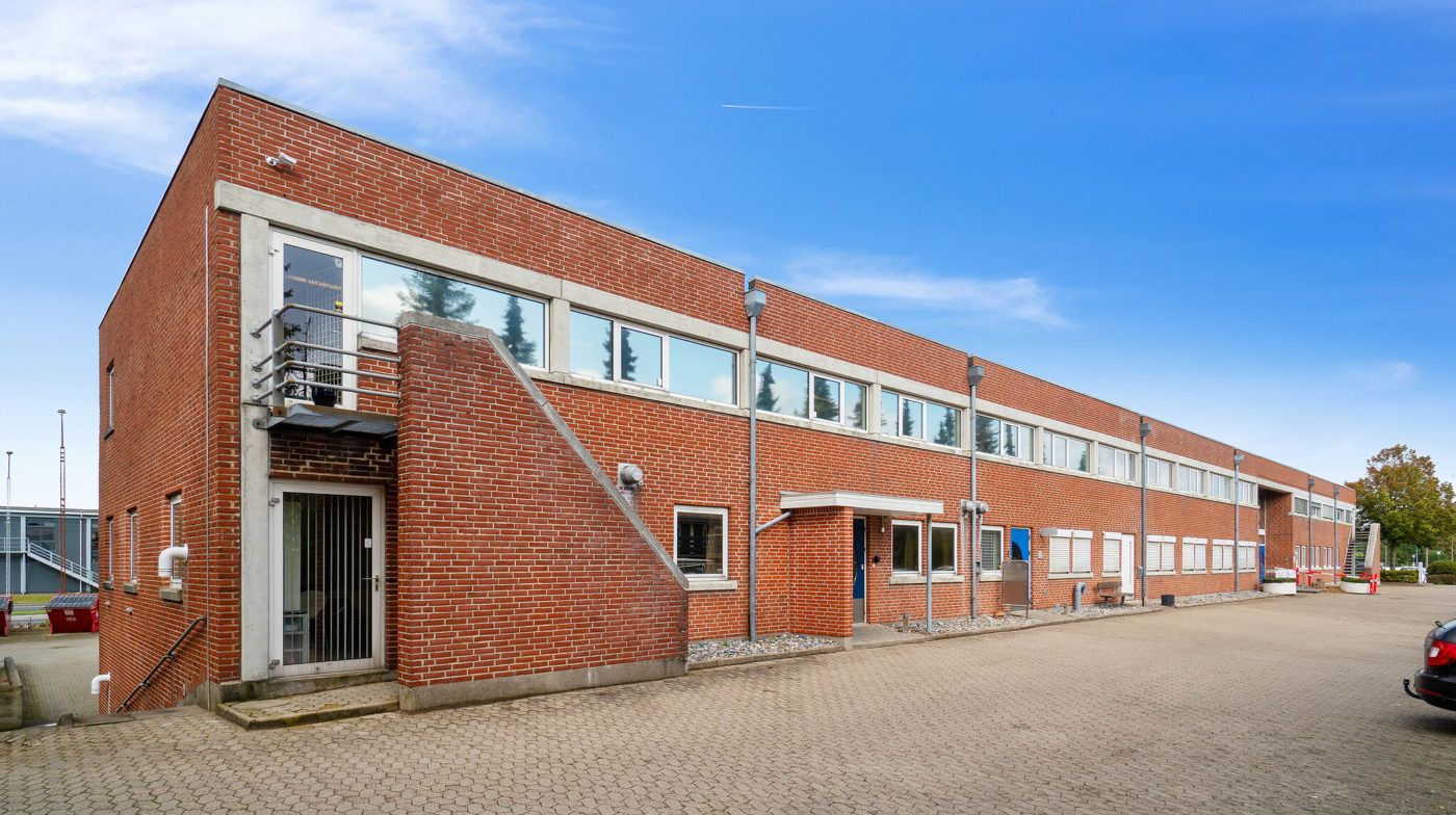 Bygning med røde mursten og store vinduer, udendørs parkeringsplads og røde affaldscontainere placeret i det fredede