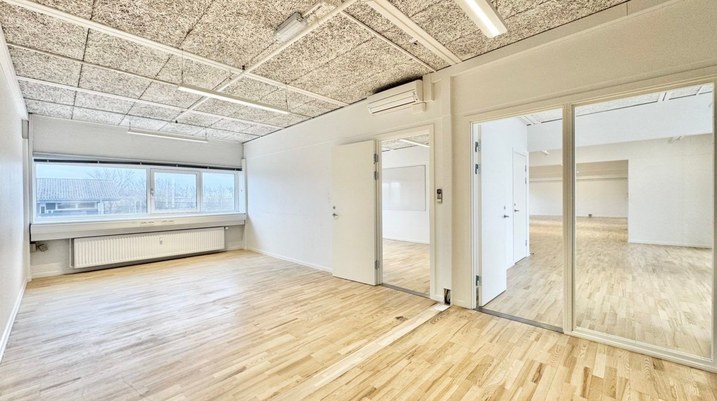 Kontorlokaler med hvide vægge og lyse trægulve samt store vinduer med udsigt til træer og hus