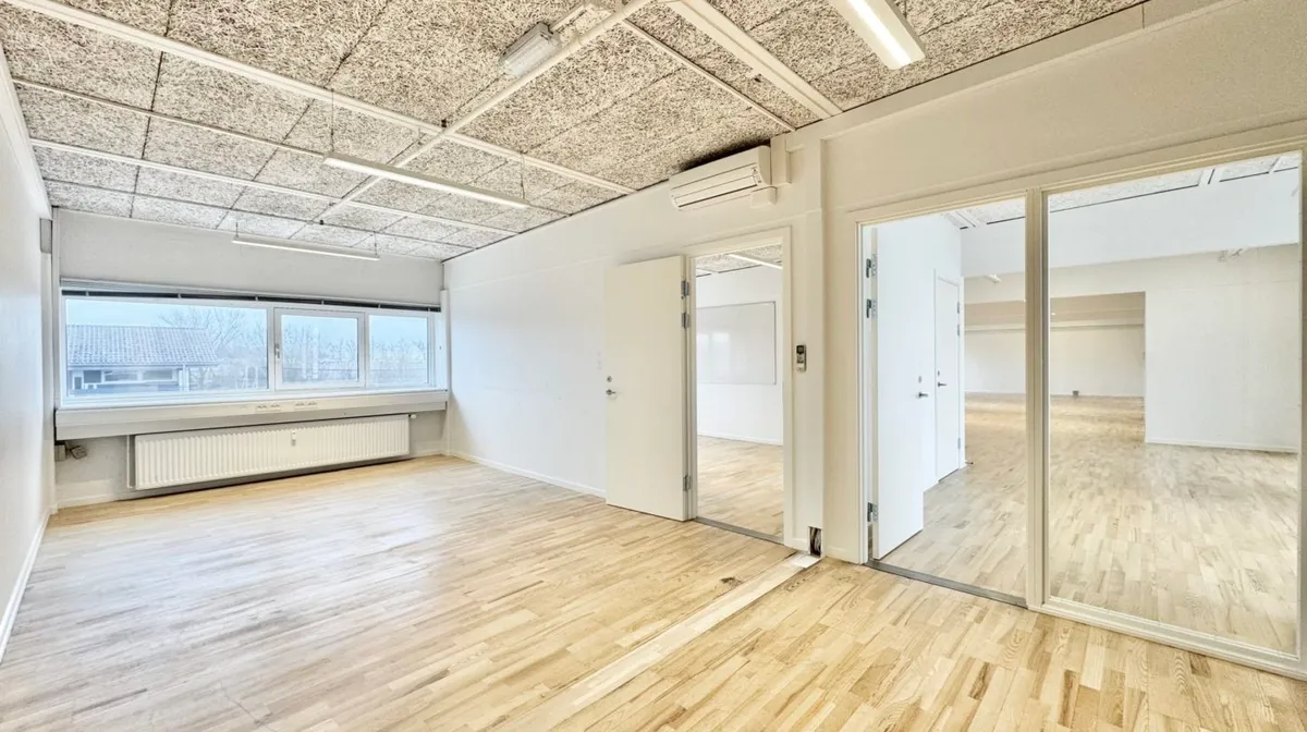 Kontorlokaler med hvide vægge og lyse trægulve samt store vinduer med udsigt til træer og hus