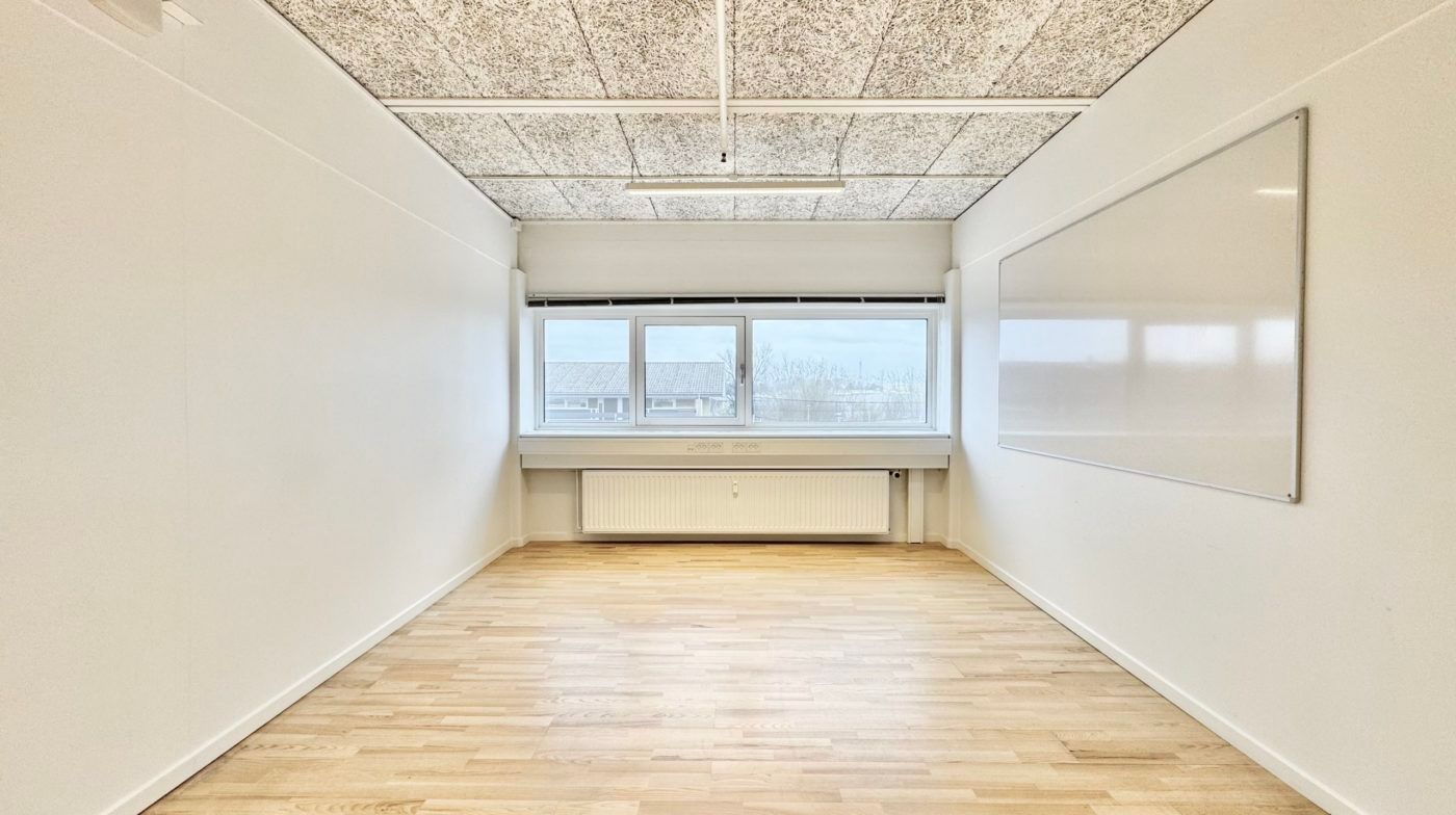 Et lukket kontorlokale med hvide vægge, en hvid tavle og et vindue med en radiator under. Loftet er lavet af gråt stof og