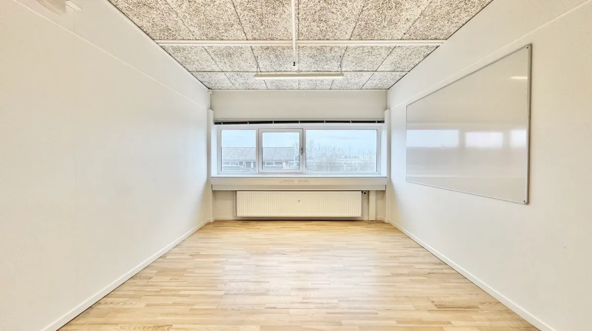 Et lukket kontorlokale med hvide vægge, en hvid tavle og et vindue med en radiator under. Loftet er lavet af gråt stof og