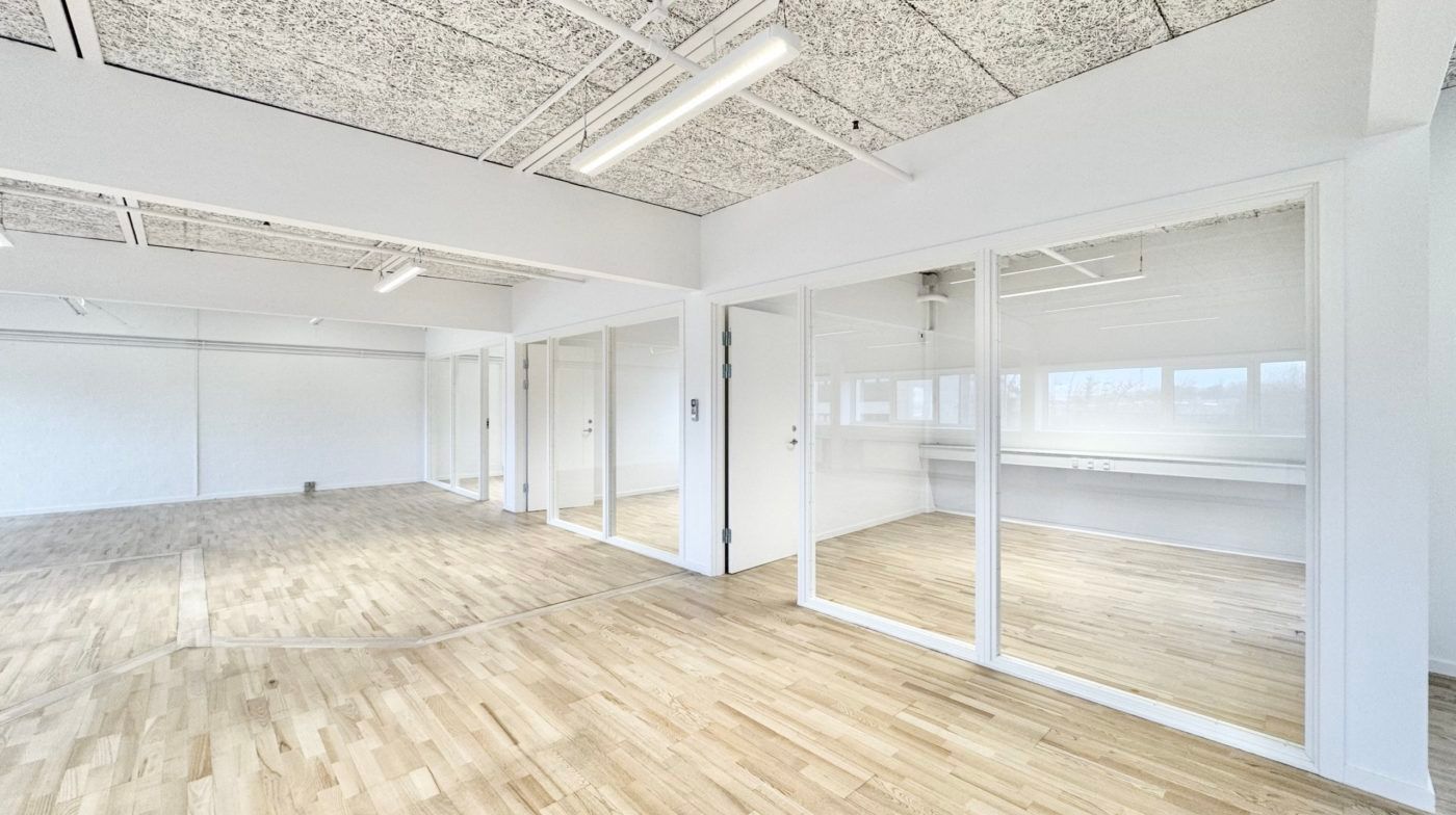 Kontorlokaler med store glasruder og højt loft i Odense med hvide vægge og lysegul trægulv samt moderne belysning