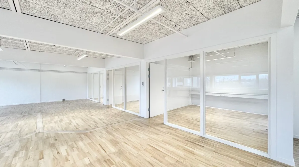 Kontorlokaler med store glasruder og højt loft i Odense med hvide vægge og lysegul trægulv samt moderne belysning