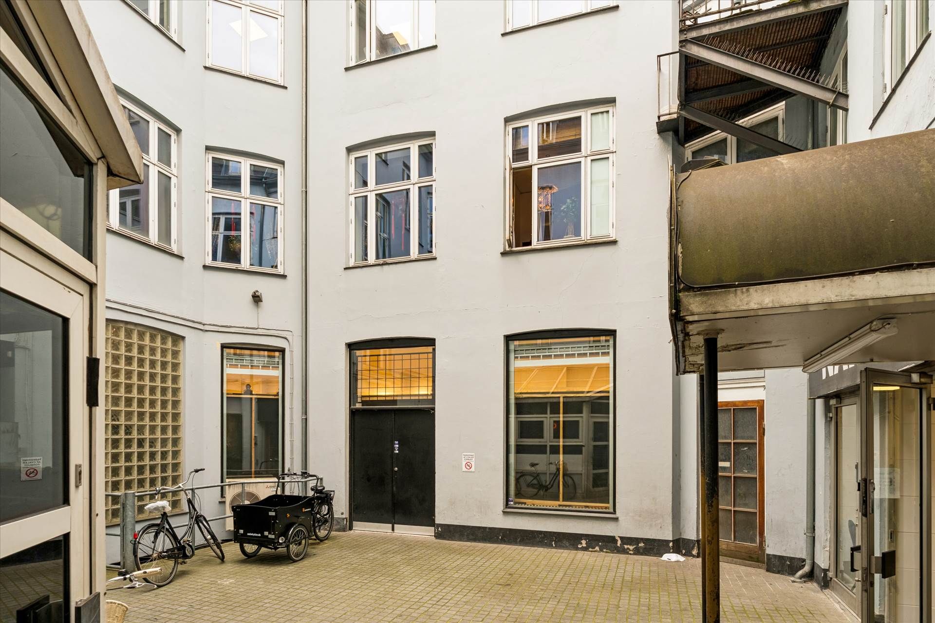 To cykler og en vogn står parkeret udenfor en hvid bygning med glasvinduer og røde døre