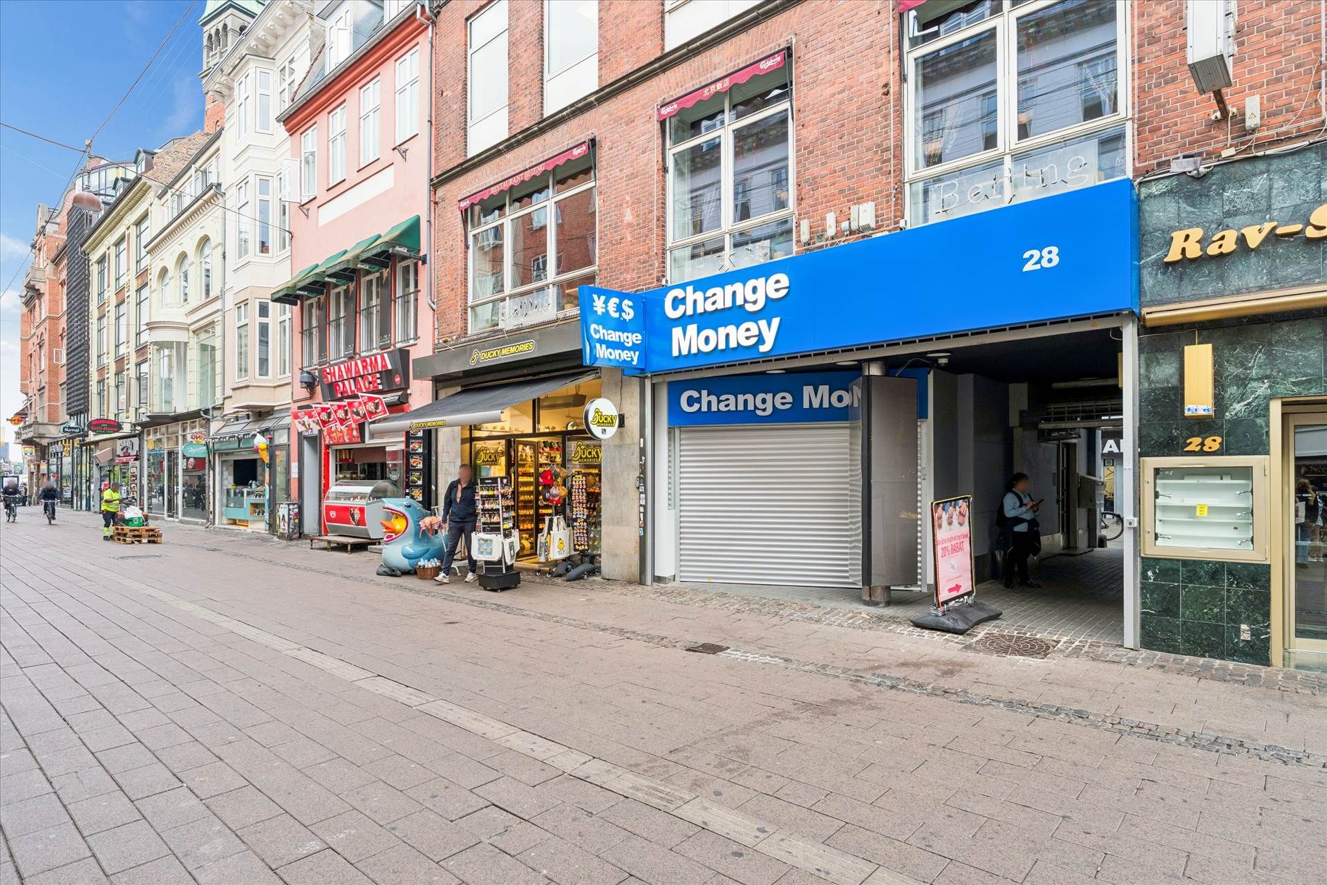 To butikker med skilte Change Money og Rav-S på en tom gade med to mænd og to cykler