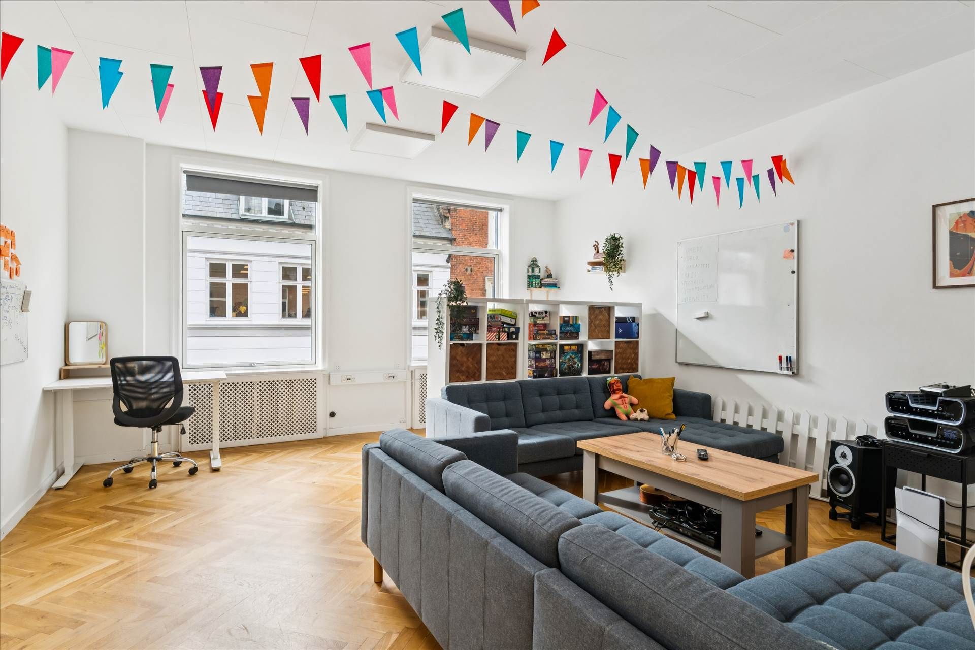 Lyst og højloftet kontor med sofaer, skrivebord, kontorstole, højtalere og minibibliotek. Farverige træflag langs loftet og