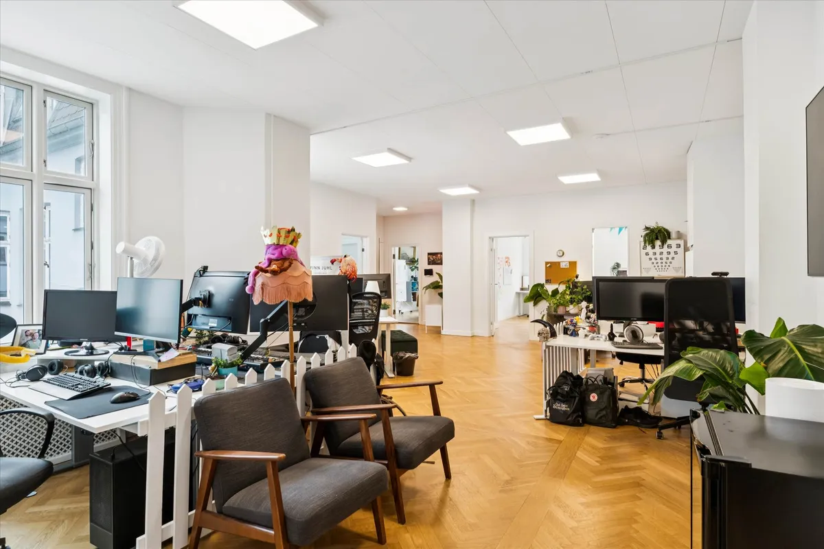 Lyst kontor med moderne møbler og højloftede værelser, fuld af skriveborde, stole, plantepoteter og computere samt et køkken