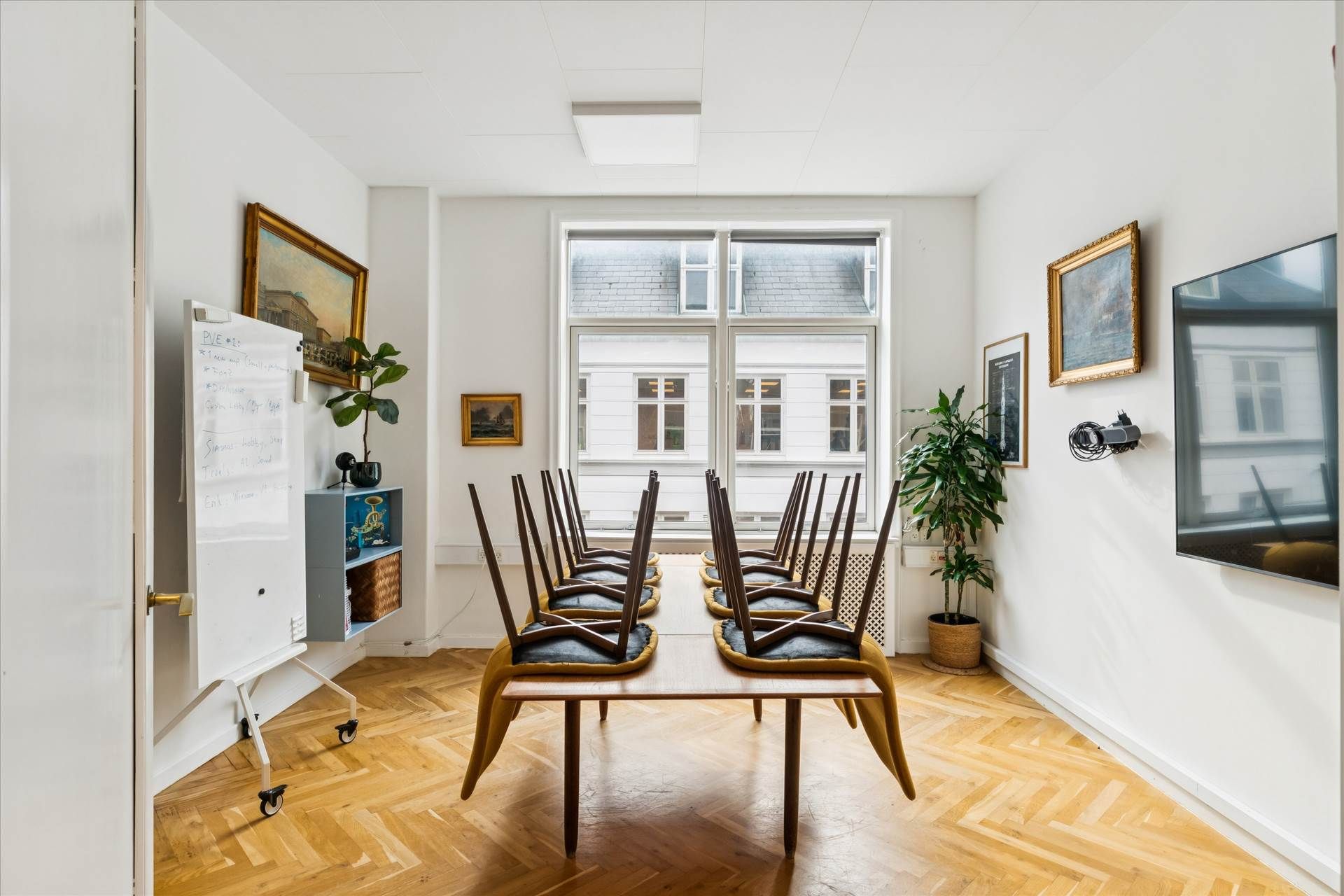 Lys kontor med trægulv og møbler. Der er en lang træbord med stole, en plante og et hvidt tavle med noter. Der er