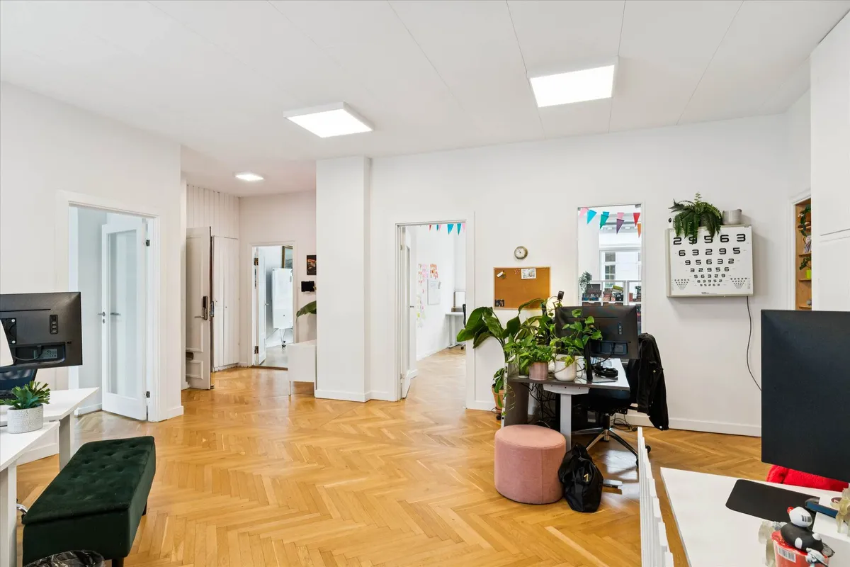 Det er et lyse kontor med højloft og trægulv. Der er et stort bord med computere og plante. Der er tæppe og sofa i en af