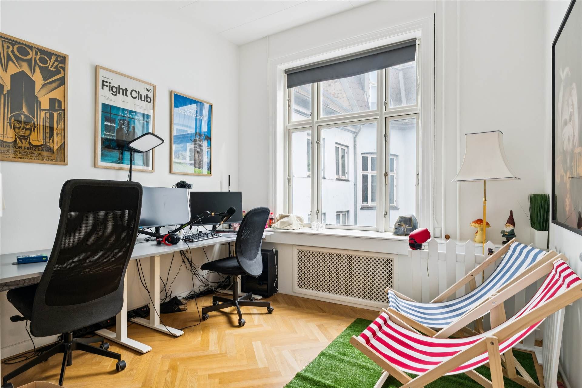 Kontor med højloft og lyse gulv. To skriveborde med skærme, hovedtøj og stole. Plakater og gardiner i baggrunden.