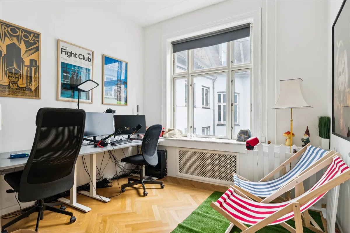 Kontor med højloft og lyse gulv. To skriveborde med skærme, hovedtøj og stole. Plakater og gardiner i baggrunden.