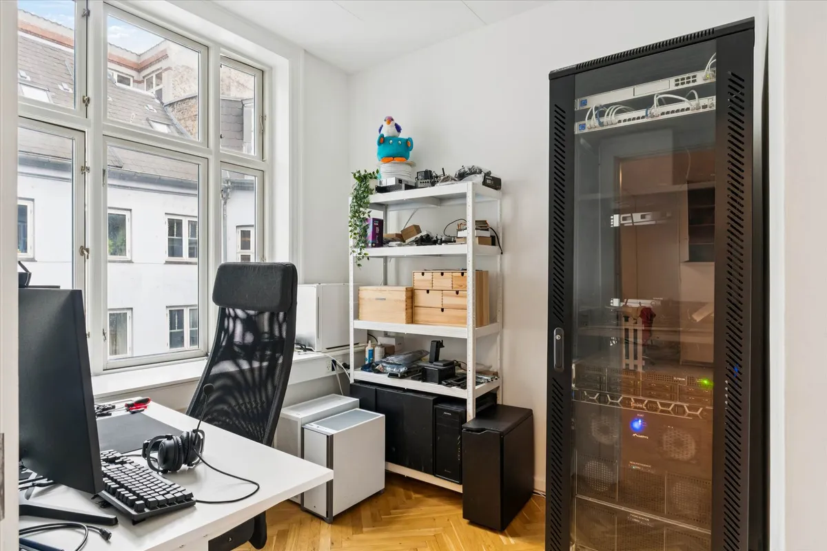 Kontor med skrivebord, computer, headset, skabe, serverskab og vinduer med udsigt til bygninger