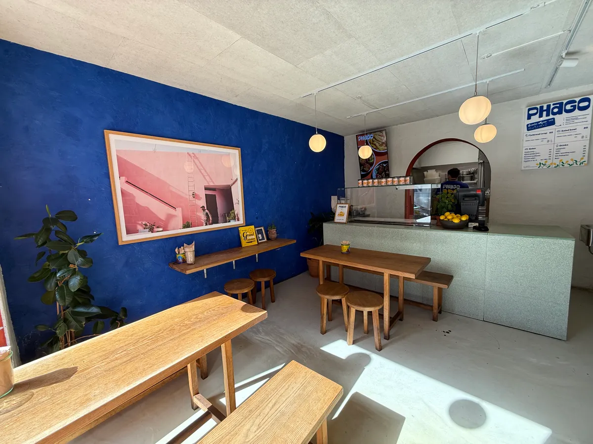 Københavns cafe med lyse og lyse træborde, smukt maleri og lyse lysekroner samt plante i hjørnet og køkken med udsigt til et