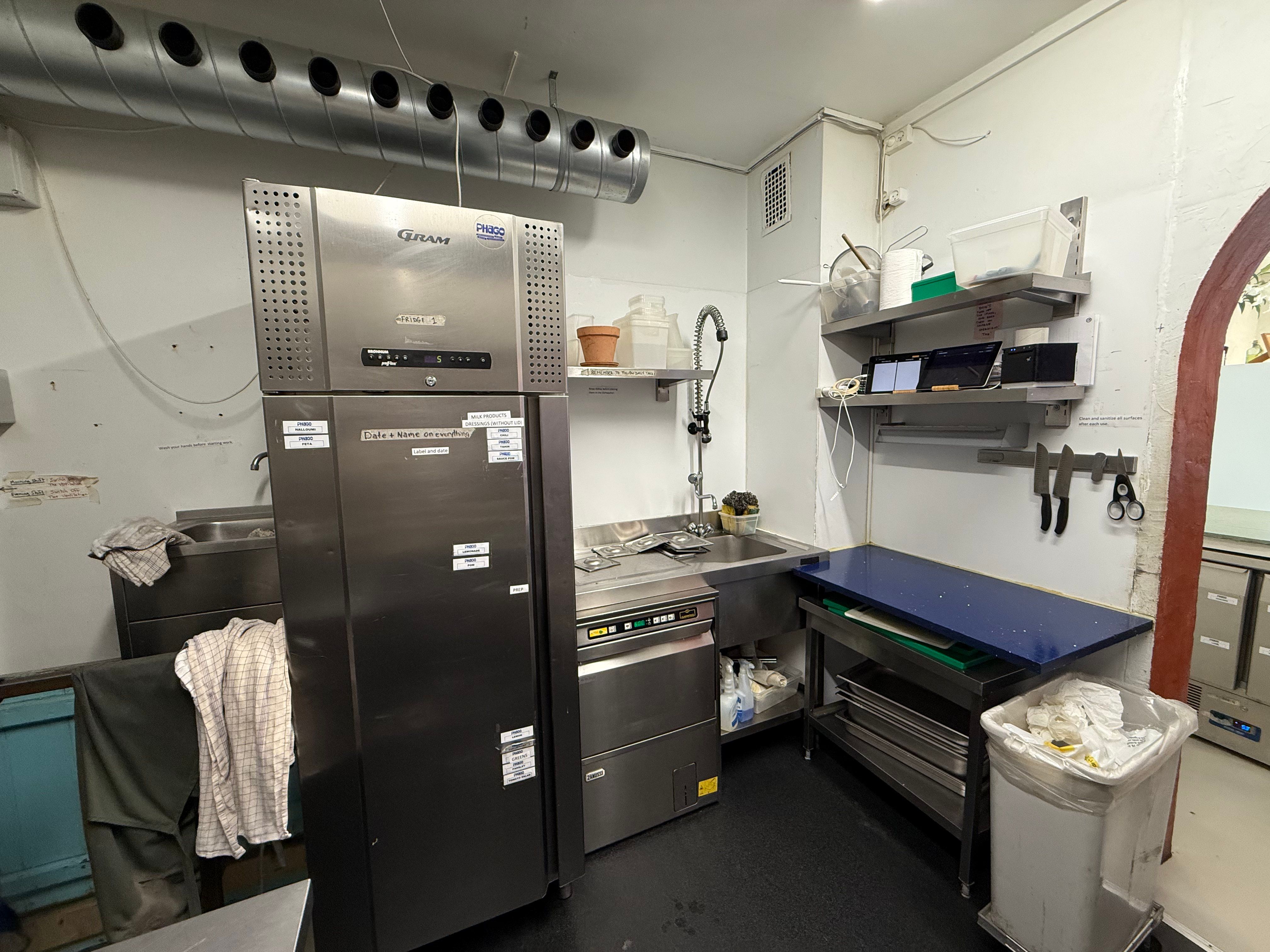 Baggrund: Et kommercielt køkken med et stort køleskab, et vaskebækken, et snitbord og en kasse med affald. Et snitbord og en