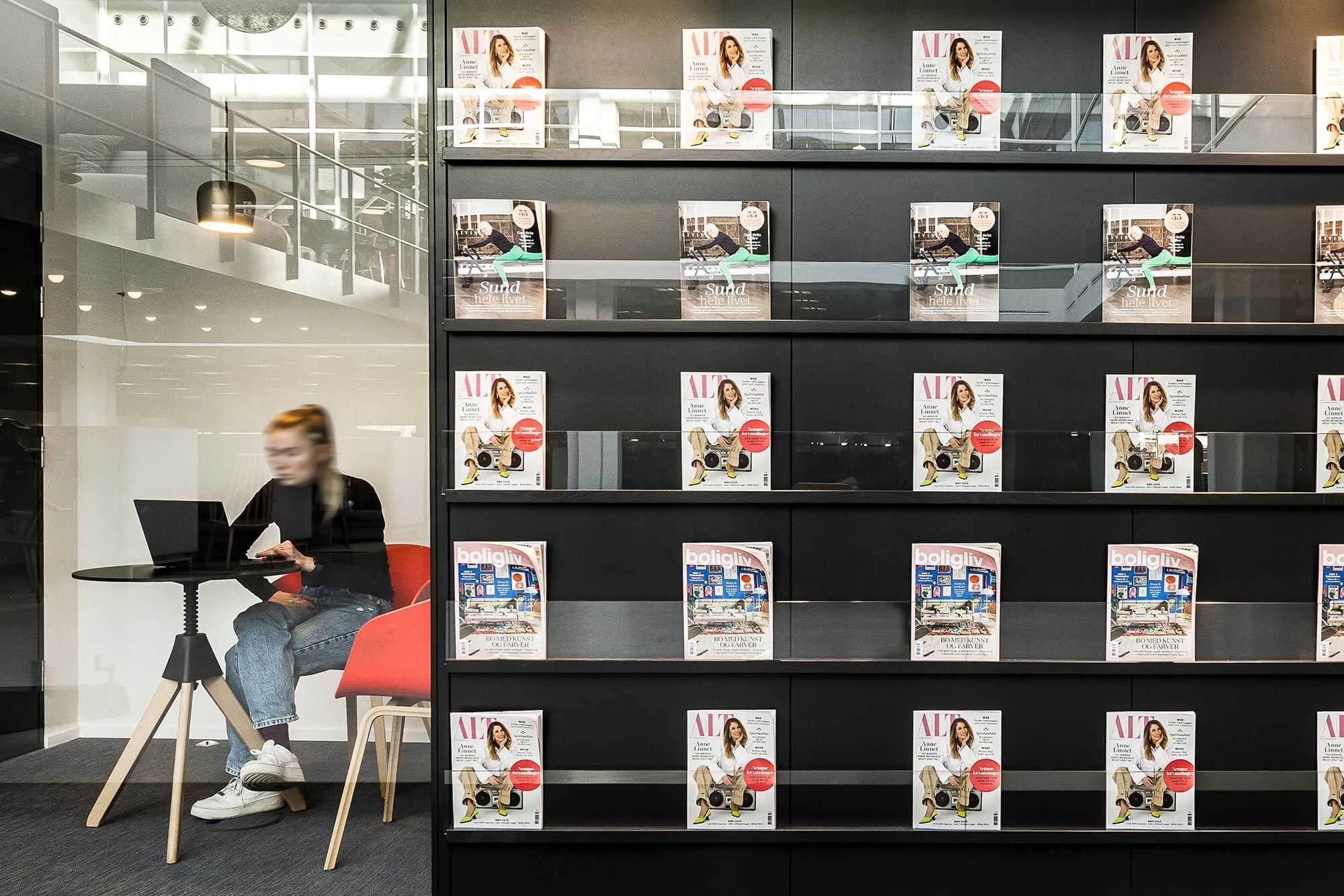 Kvinde arbejder med bærbar computer på kontor med glasvægge og moderne møbler og en hylde med tidsskrifter