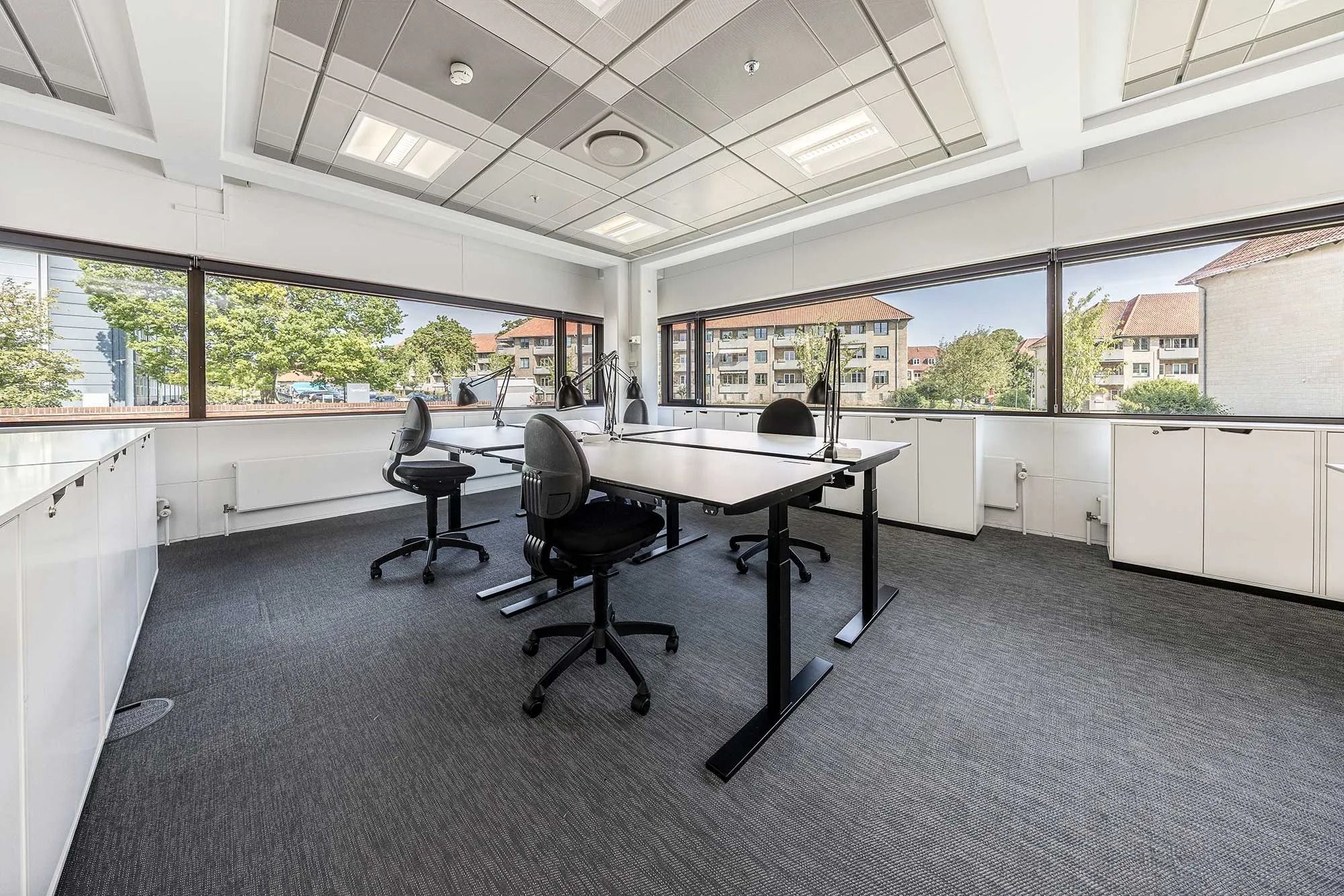 Kontor med høje ruder og lyse lofter, hvor skriveborde med ergonomiske stole står. Højteknologiske møbler og lyse vægge