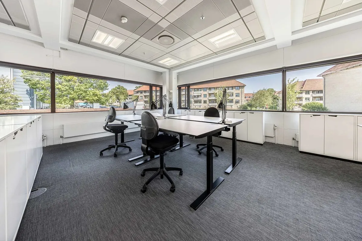 Kontor med høje ruder og lyse lofter, hvor skriveborde med ergonomiske stole står. Højteknologiske møbler og lyse vægge