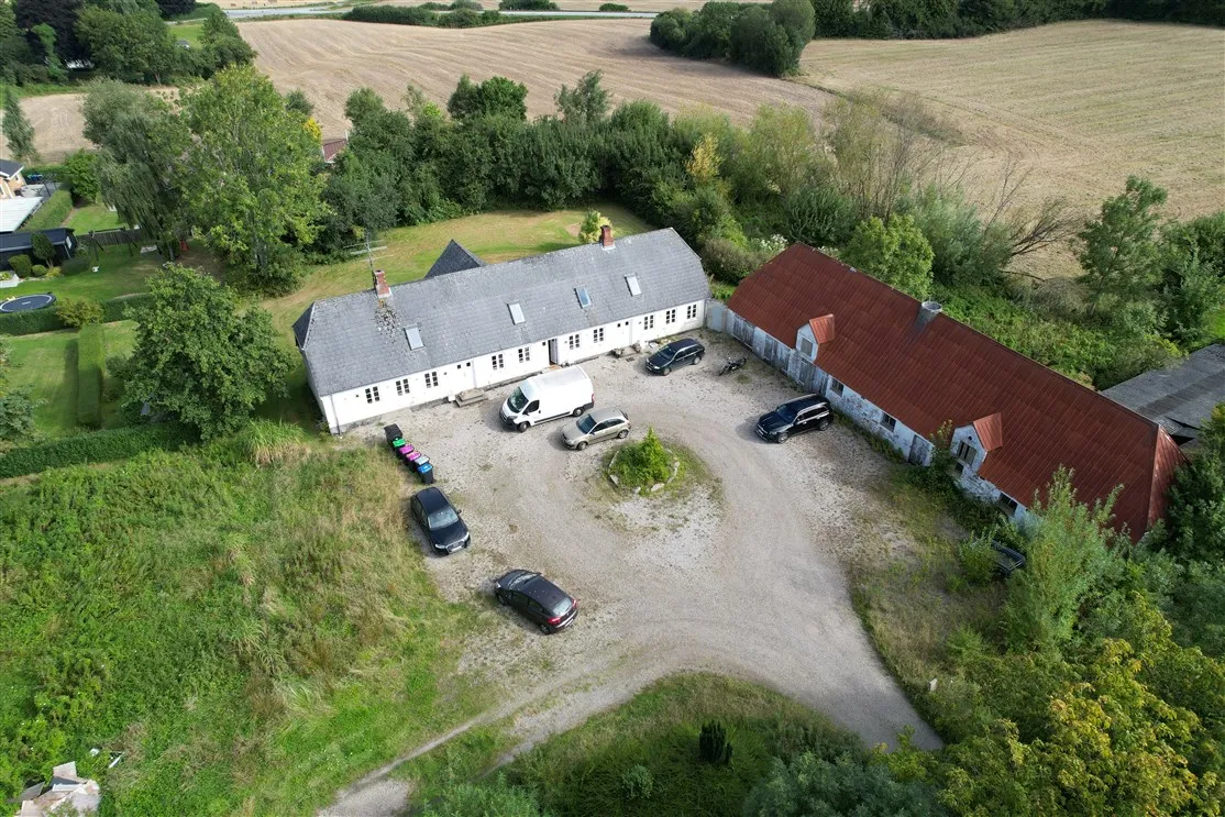 Flere biler parkeret foran en stor bygning med grå tag, samt en række mindre bygninger i baggrunden