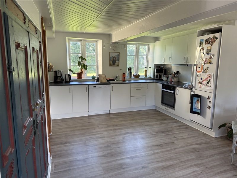 Køkken med hvide vægge og trægulv, hvidt køkken med ovn og køleskab, små træbord med køkkenudstyr og plante