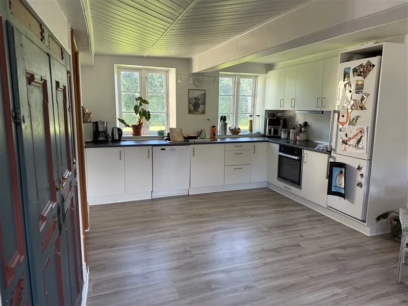 Køkken med hvide vægge og trægulv, hvidt køkken med ovn og køleskab, små træbord med køkkenudstyr og plante