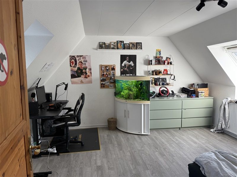 Et værelse med et skrivebord, en computer, en stol og en fiskestand samt et køleskab og et vindue i væggen