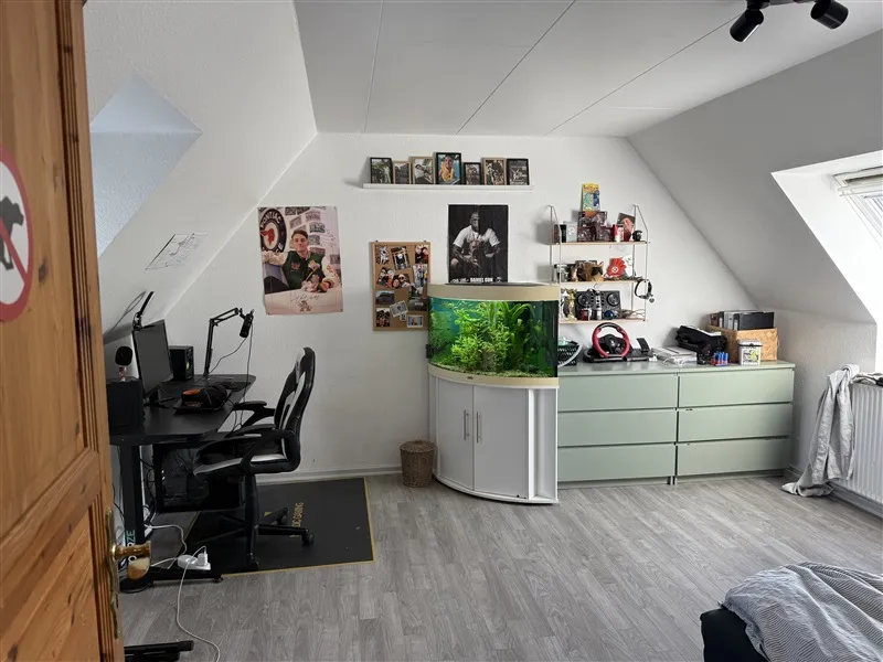 Et værelse med et skrivebord, en computer, en stol og en fiskestand samt et køleskab og et vindue i væggen