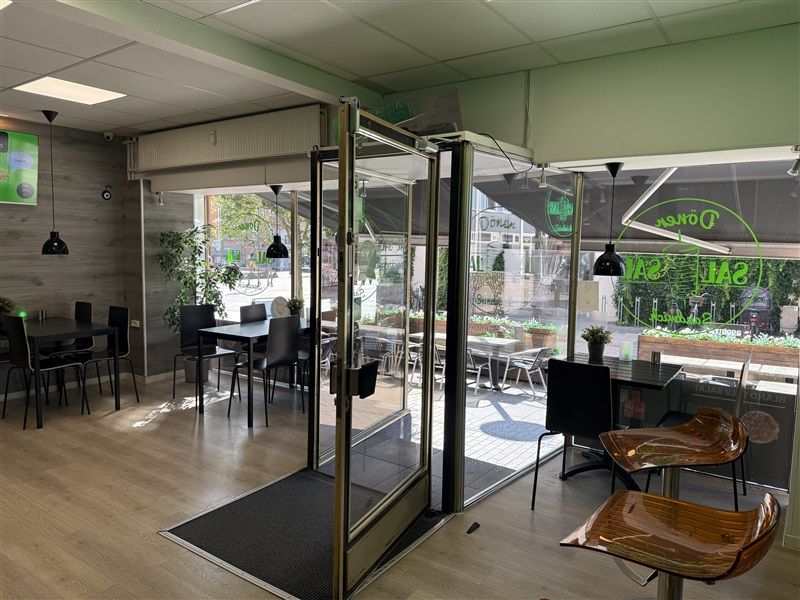 En møbleret café med indgangsdøre, vinduer og trægulve, samt træborde og stole samt plante i plastikpotte