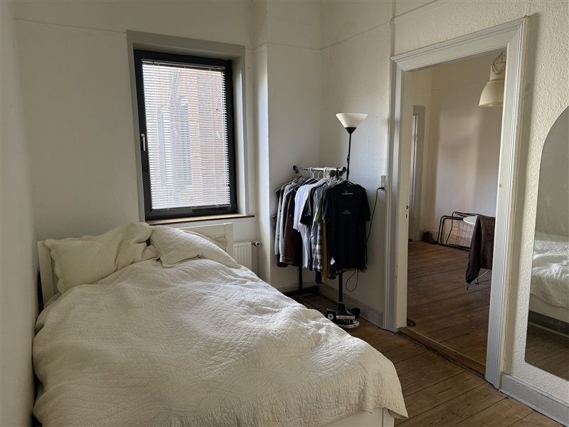 Et soveværelse med et dobbeltseng, to puder og en tøjstativ med tøj samt en lampe og et vindue med gardiner