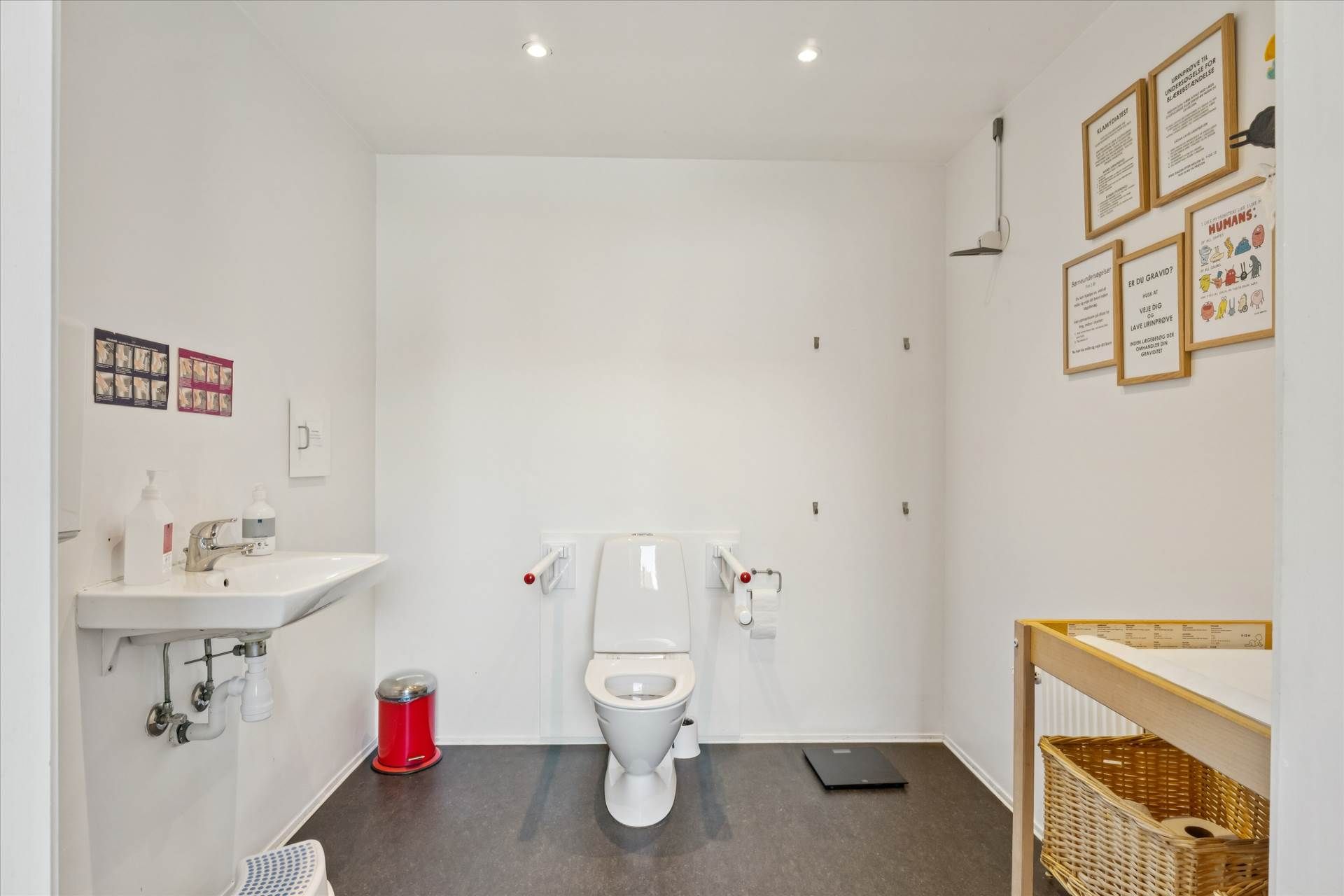 Et hvidt toilet med toiletpapirhylde og håndklædetørrer. Hjørne med vandhane og håndfat samt et toilet med hjælpegreb. En