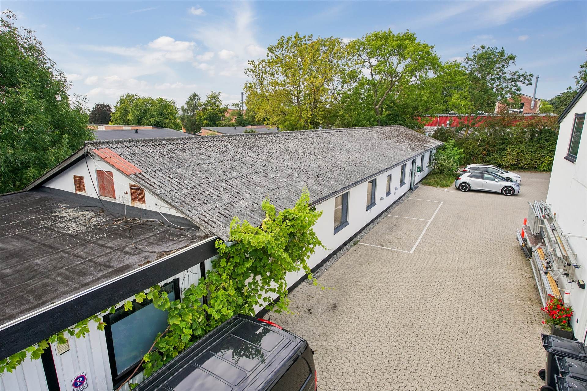 En række hvide erhvervsejendomme med parkeringsplads og træer samt et parkeret elektro- og et dieselbil.