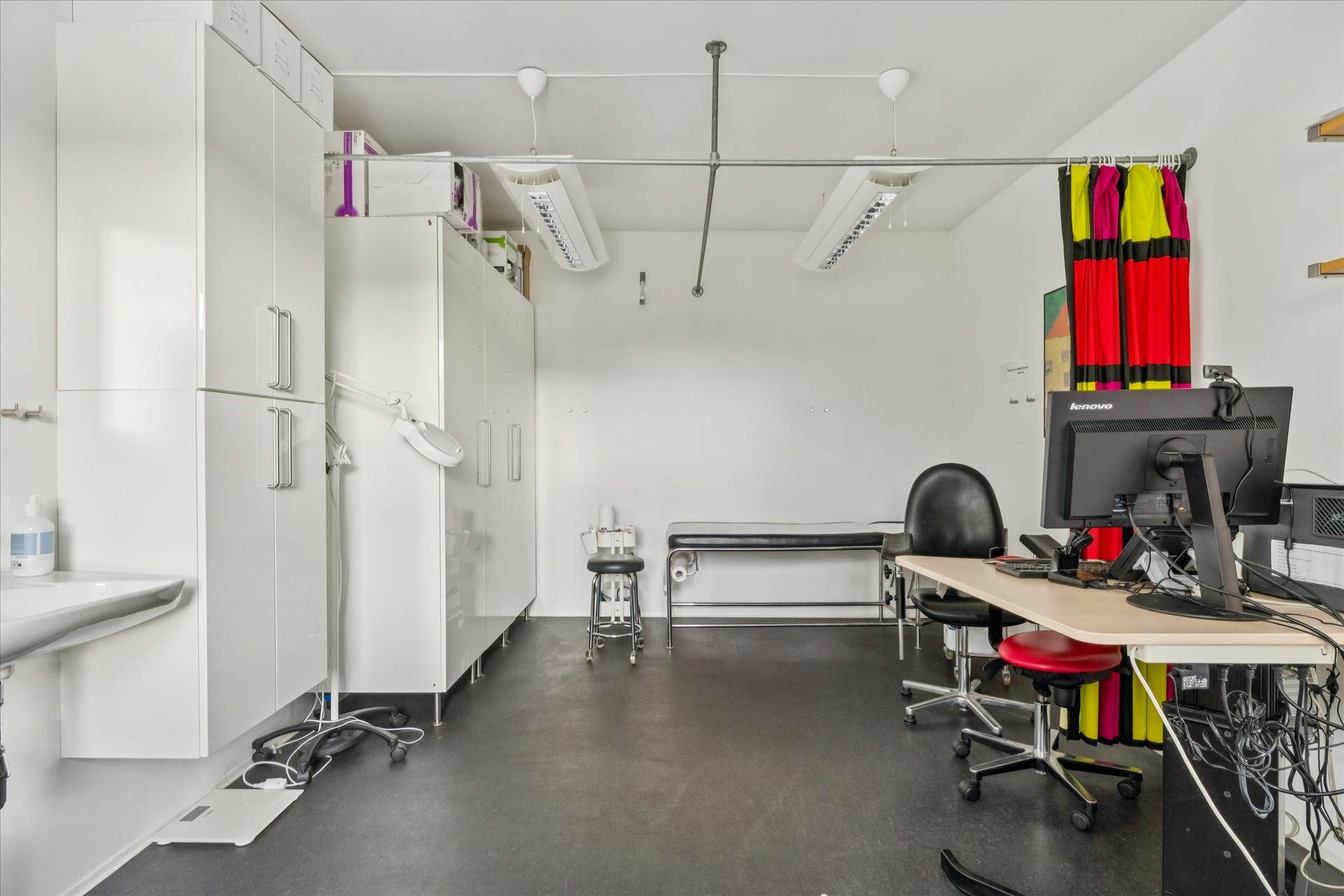 Lægeværelse med højloft og hvide vægge, hvor der er monteret to lysekroner, hvidt skab med håndtag, toilet, computer, og