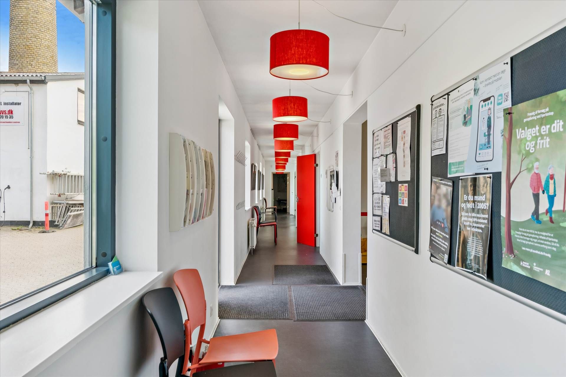 Lang korridor med røde lampeskibe og lyse hvide vægge, samt lægehusrelaterede plakater og annoncer