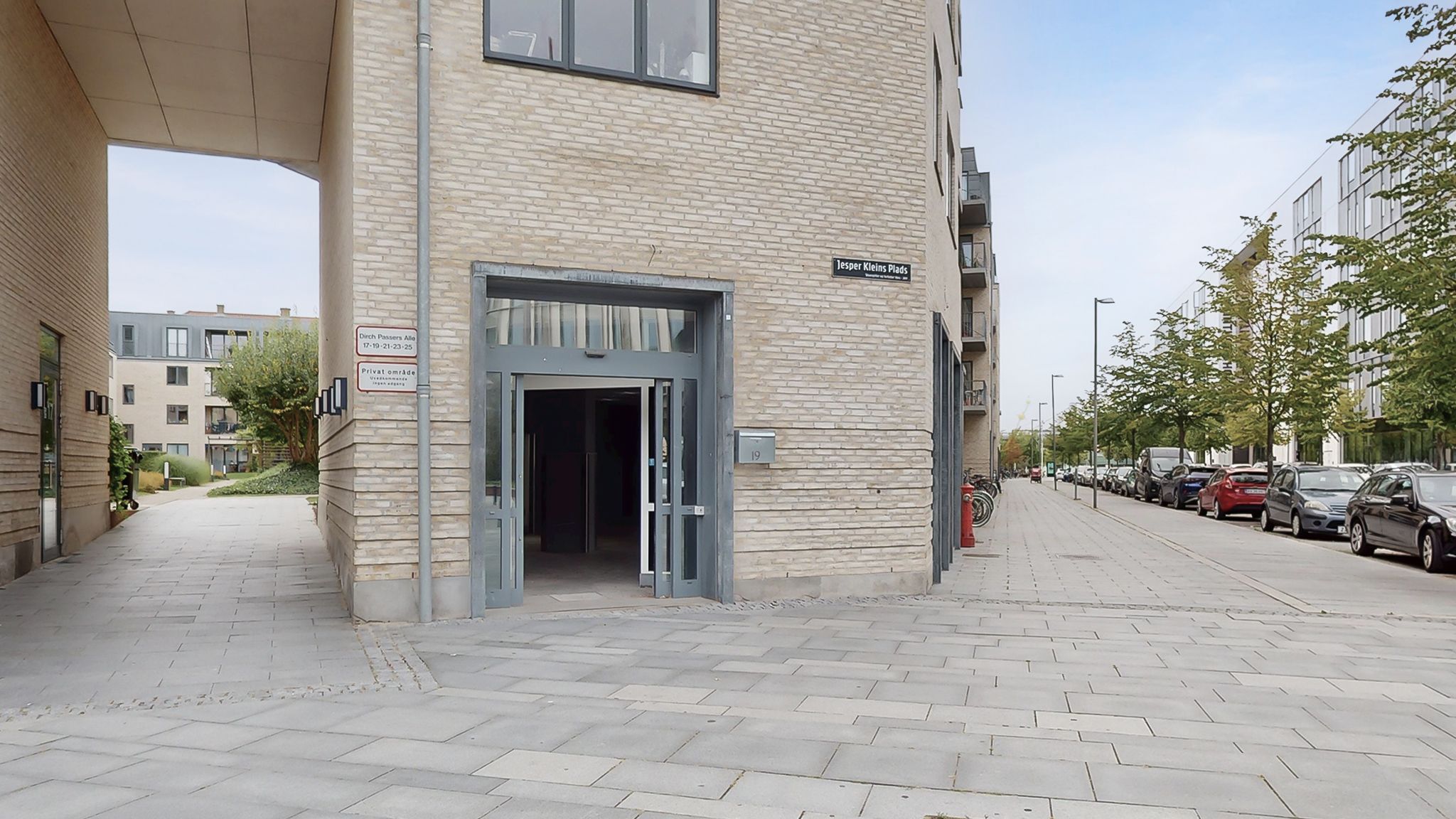 En slank stenbygning med et åbent indgangsparti og en søjle med plaketterne "Dirch Passers Alle" og "Jesper Kleins Plads" på