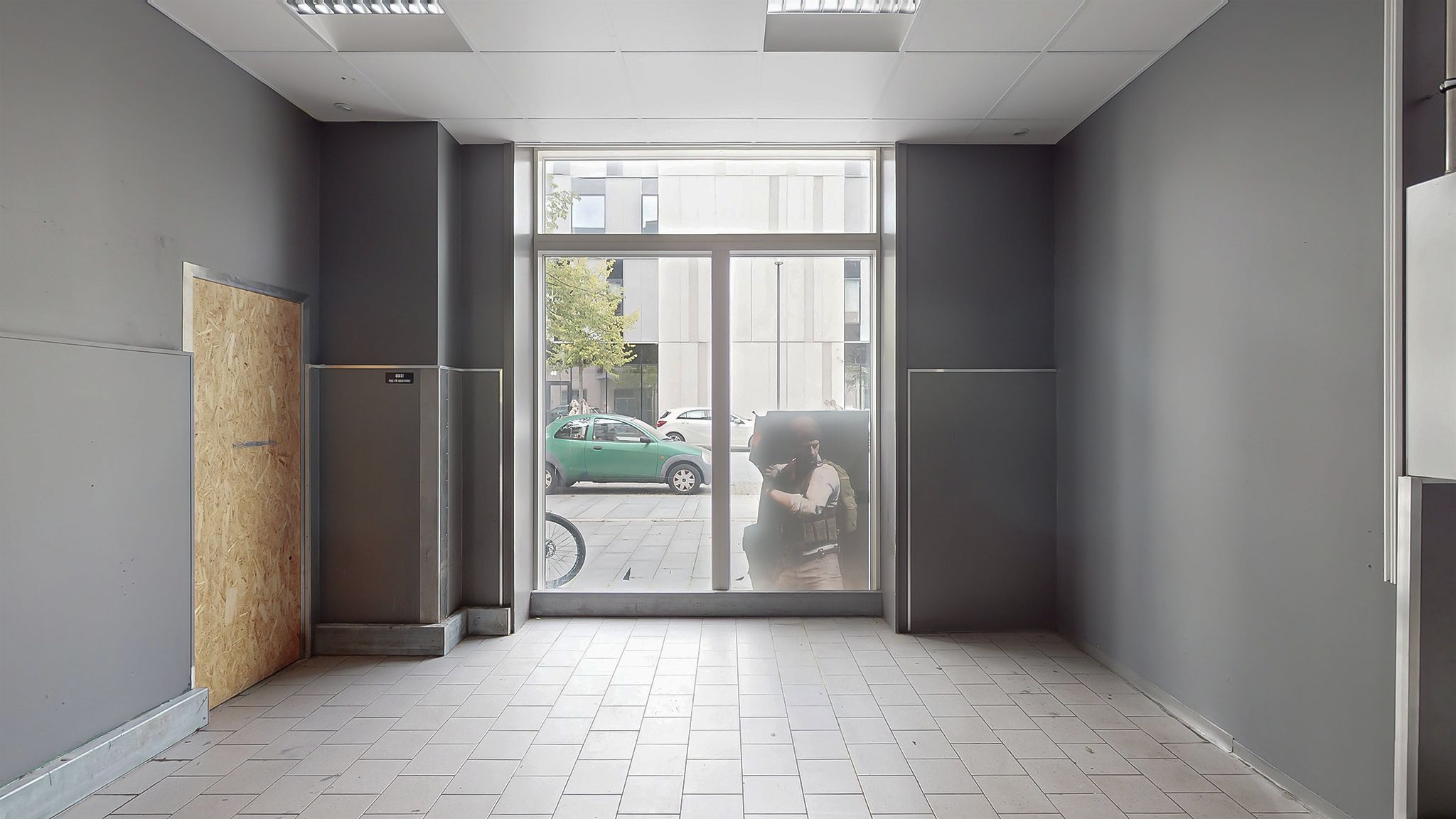 Et lejlighedslokal med hvide flisegulve og lysegrå vægge. Der er glasdøre og en åben dør med træbeklædning. En cykel står