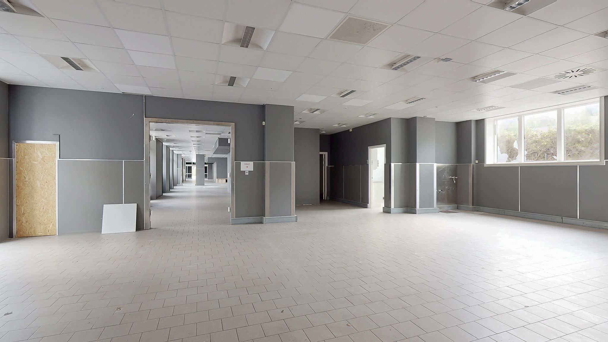 Lang, tom gang med hvide gulvfliser og høje, lyse vægge. Adgangsdøre og skraldespande står i gangen. Et vindue viser ud til