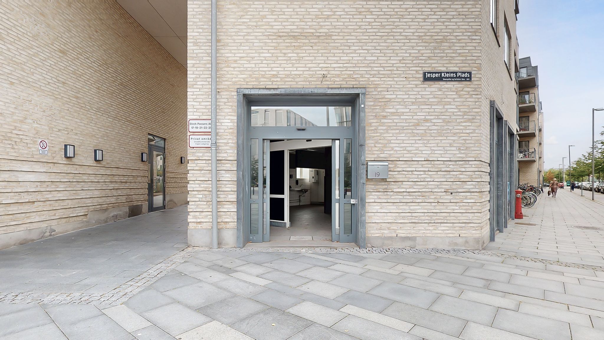 En moderne bygning med åbne døre og indgangsområde, hvor der er et skilt med Jesper Kleins Plads. Bag indgangen er der et
