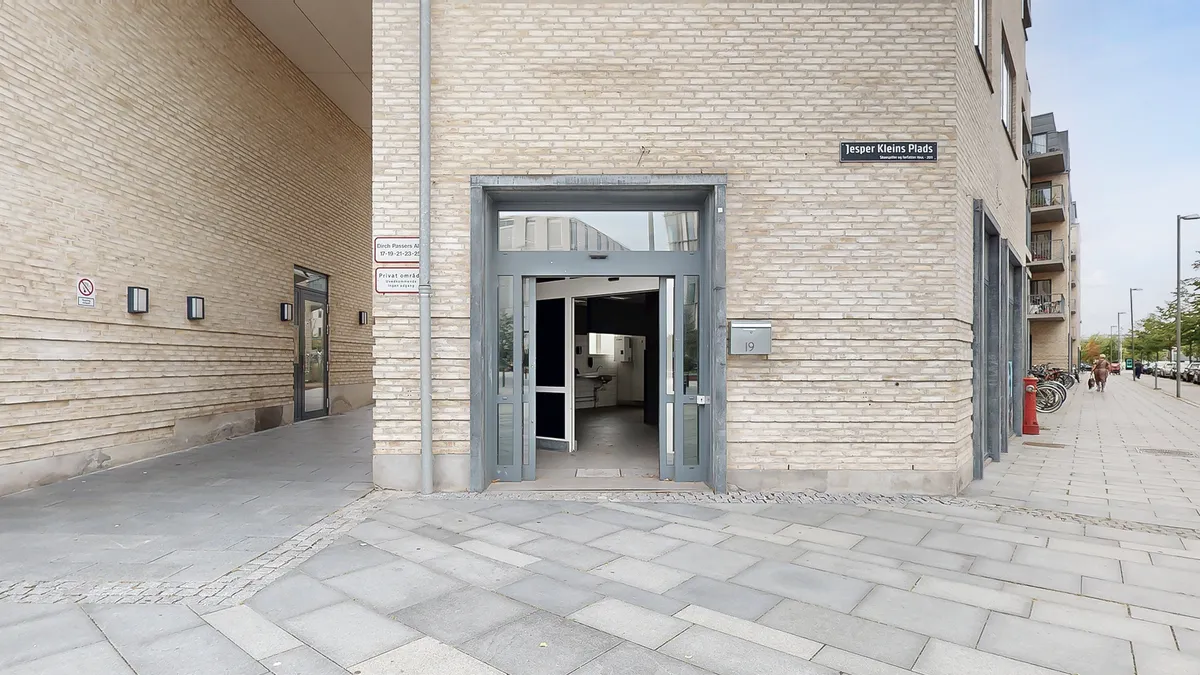 En moderne bygning med åbne døre og indgangsområde, hvor der er et skilt med Jesper Kleins Plads. Bag indgangen er der et