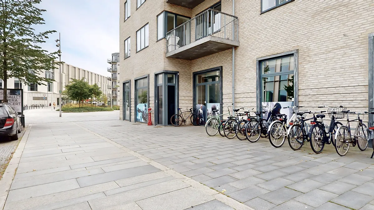 En bred gågade med parkerede cykler foran et lejlighedsbyggeri med en indgang og et højt vindue