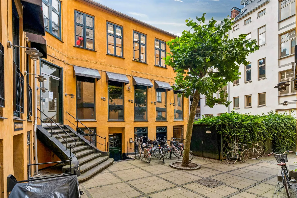 Kort række med parkerede cykler og stigning i gult bygningshus, udsigt mod en stor busket og et træ