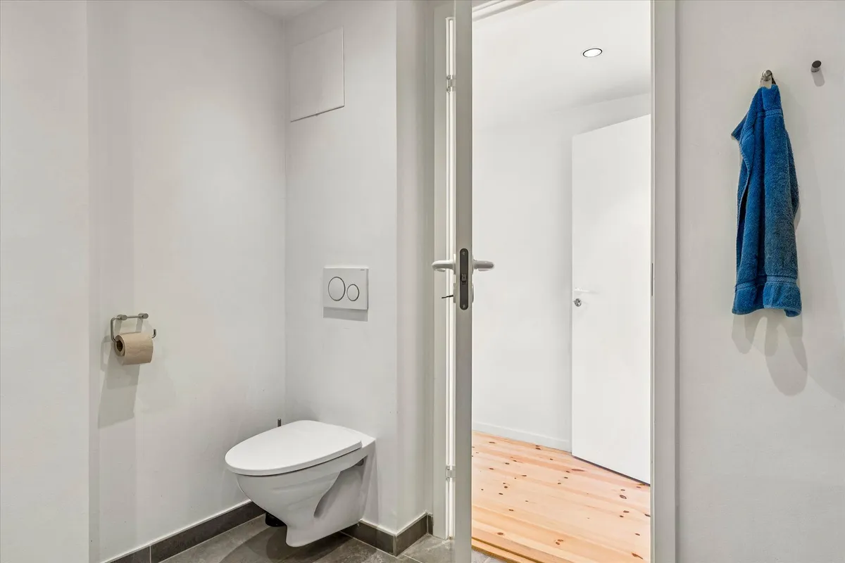 Et lille, moderne og rent badeværelse med et toilet, hvor en hvid dør er åben og en blå håndklæde hænger på væggen.