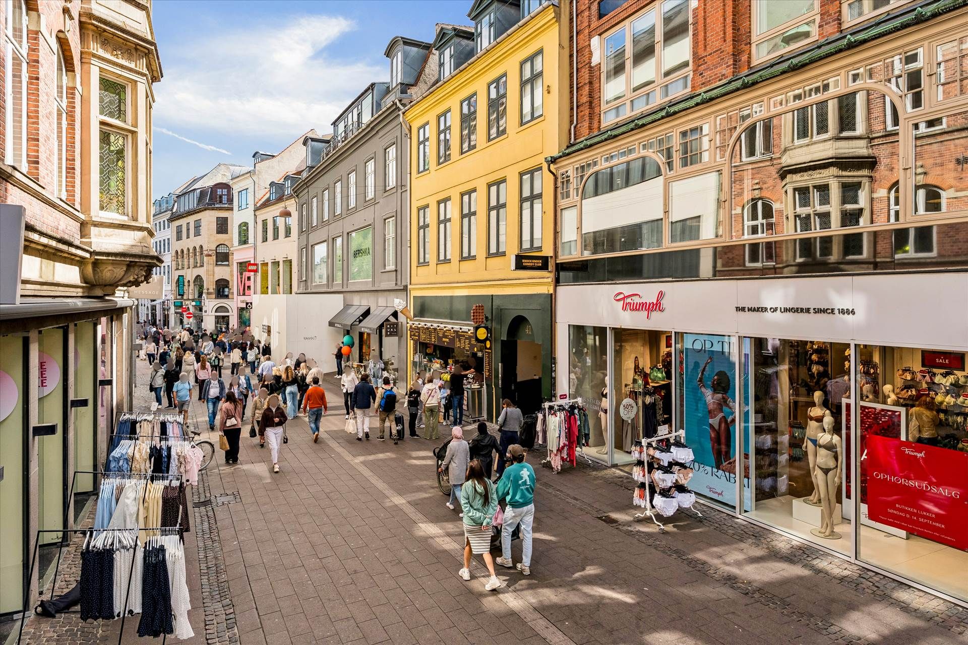 Mennesker vandrer rundt på en gågade i København, der er fyldt med butikker og mærkeskilte, og folk går forbi en række