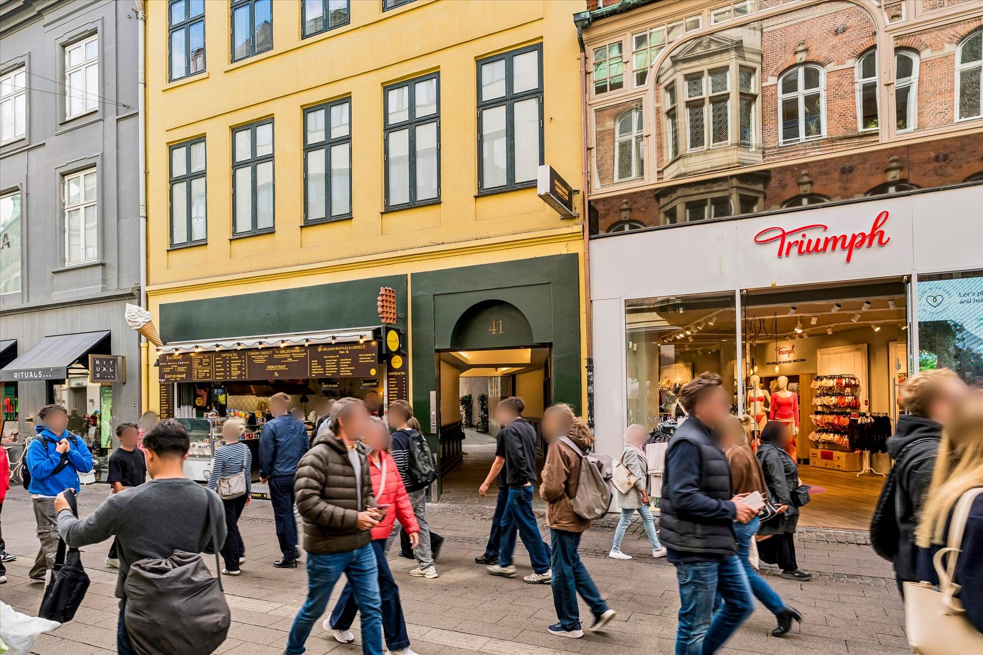 En række mennesker går på en gågade foran en Triumph-butik og en isvogn på Vimmelskaftet i København