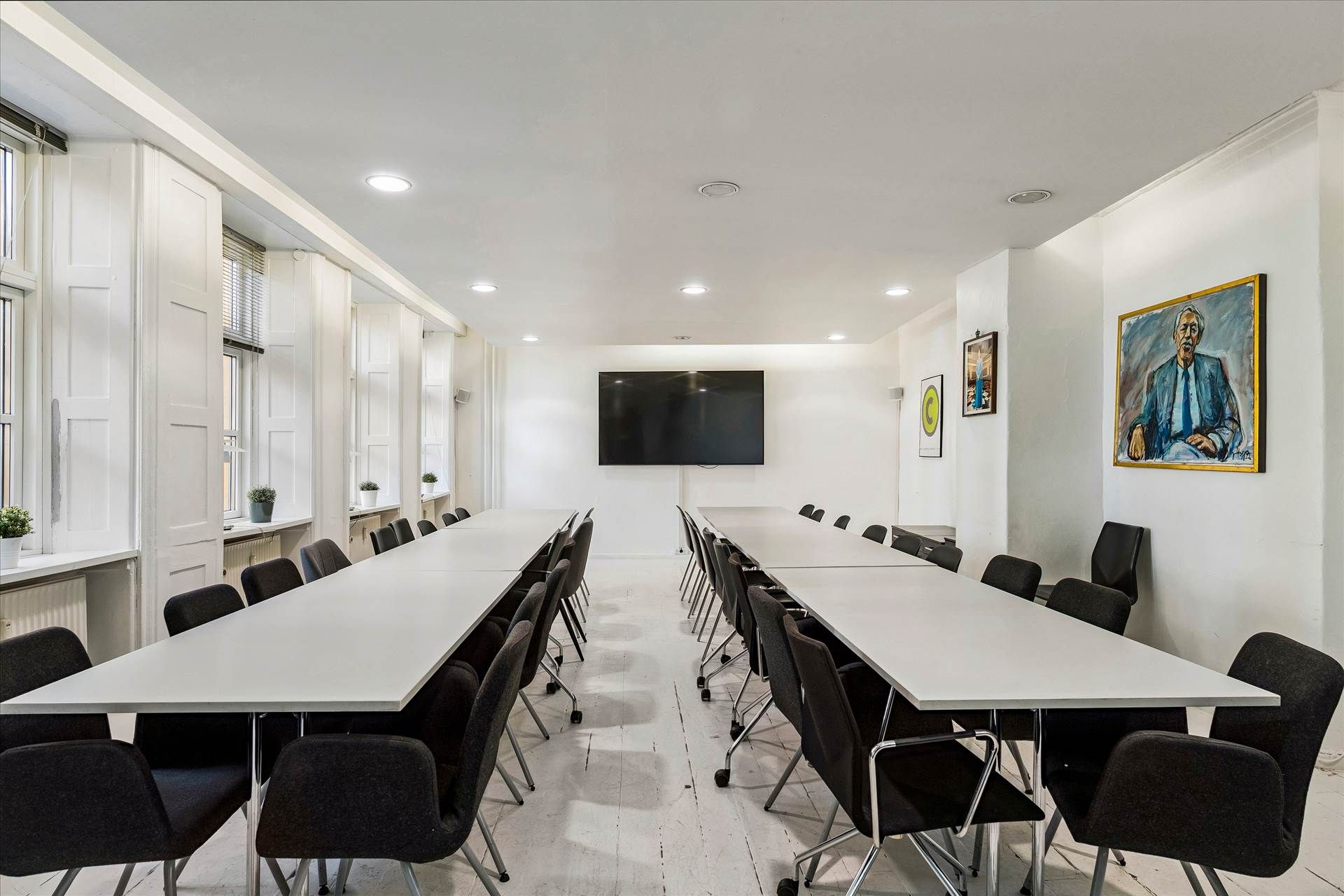 Moderne konferencesal med hvide vægge og trægulve, hvor fire lange borde er dækket med mørke stolen og en stor tv-skærm er