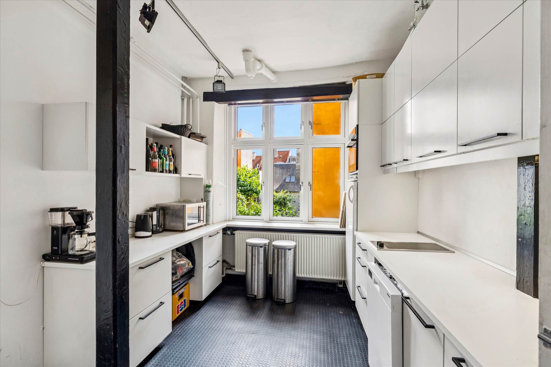 Et moderne køkken med hvide vægge og højloft, to høje højstander og to stålskraldespande og en mikrobølgeovn på en bordplade.