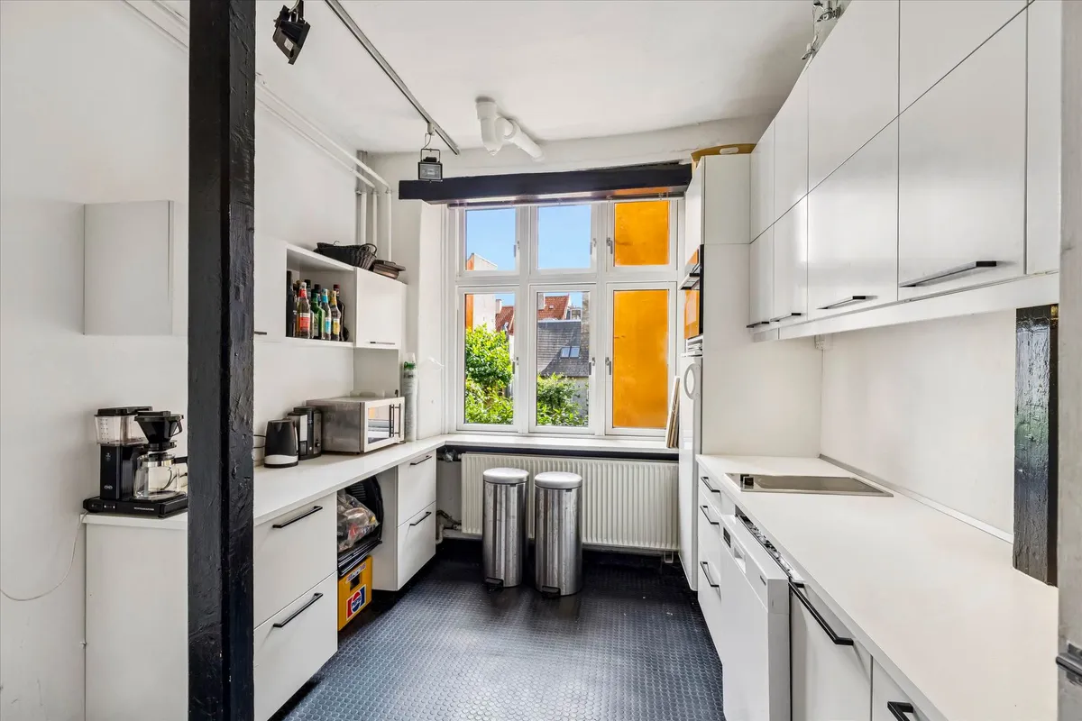 Et moderne køkken med hvide vægge og højloft, to høje højstander og to stålskraldespande og en mikrobølgeovn på en bordplade.