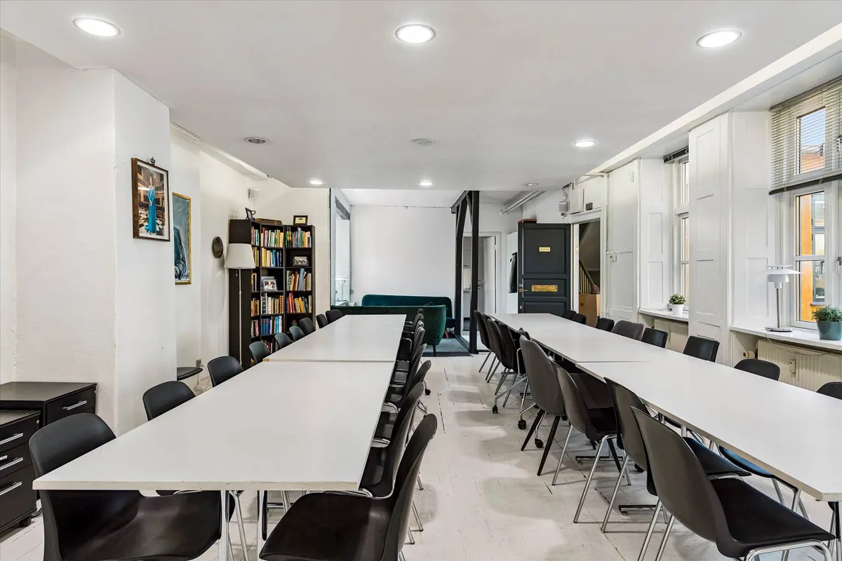 Lys, moderne kontor med lange borde og stole, højt til loftet, gulve og hvide vægge, bibliotek med bøger, sofa og køkken.