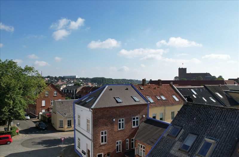 Byudsigt med røde og gule huse, røde tag, lyseblå himmel med skyer og en røde bilparkeret parkeringsplads
