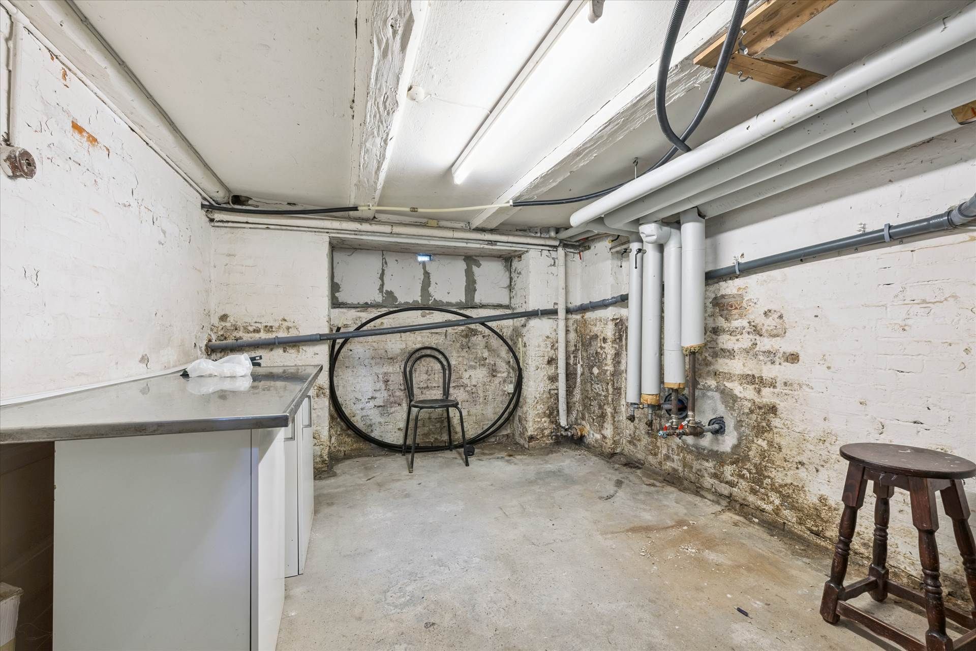 En værkstedslokal med højt til loftet, træstol, et bord med en plastikpose og en stol i et mørkt hjørne med rør og røgkammer